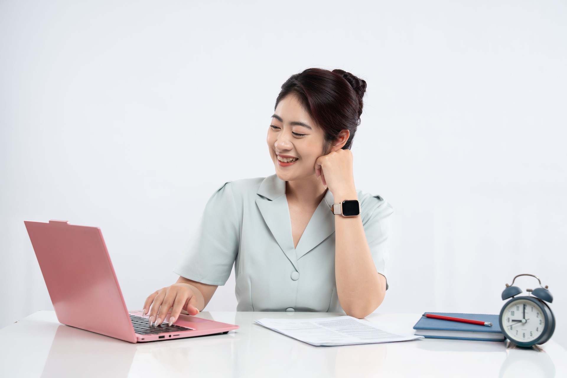 Woman working at a desk using a laptop with an alarm clock notebook and pencil nearby